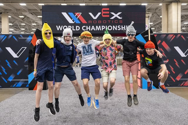 A V5RC team wearing an assortment of silly hats jumps together beneath a VEX Robotics World Championship banner.