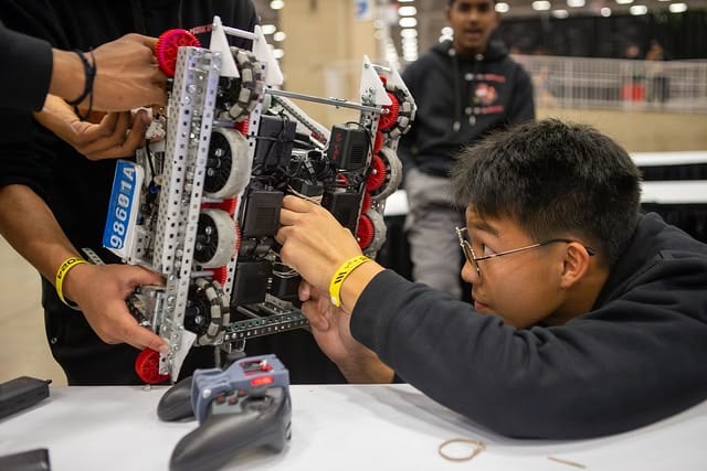 로봇공학 전공 학생이 경쟁 로봇의 아랫부분에 있는 모터를 조정하는 동안, 다른 두 팀원이 로봇을 들어올리고 있습니다. 그들 앞 테이블 위에는 통제자가 앉아 있다.