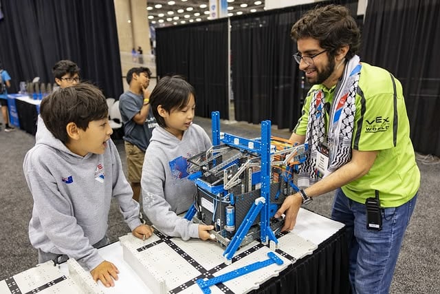 Two members of a robotics team talk with a volunteer at the inspection table, as the volunteer uses the VEX IQ sizing tool to make sure the robot is the correct size for competition.