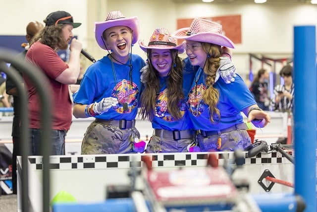 Three girls on a robotics team in matching tshirts and pink cowboy hats are hugging and laughing beside a game field, while an announcer speaks into a microphone behind them.