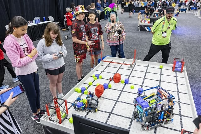 Two teams stand beside a VIQRC Field playing a qualifying match at the 2024 VEX Robotics World Championship. There are two robots on the field picking up and scoring blocks, while a referee looks on and an emcee announces the match into a microphone. 