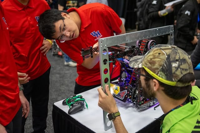 A V5RC team in red shirts stands holding part of their robot, while a volunteer inspector holds up a V5RC inspection measuring took to ensure the robot is within the acceptable size limit on the inspection table.