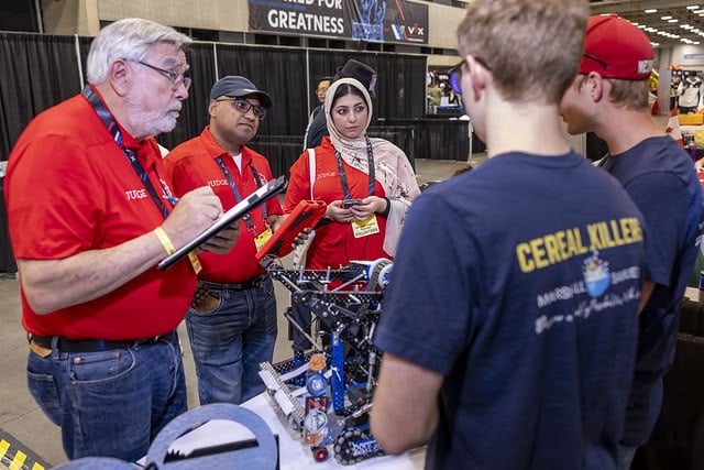 Three judges in red shirts stand talking to members of a V5RC team about their robot, placed on the center of the table between the two groups.