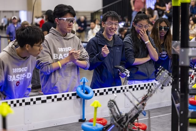 Two teams stand beside a field during a match with various expressions as they cheer, drive, and compete together as an alliance in a qualifying match.