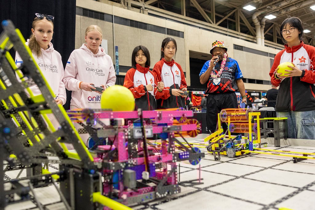 Two VIQRC Robotics Teams concentrate on a match at the VEX World Championships. The team on the left is holding a controller as their robot prepares to shoot a ball into a goal. The team on the right observes, while one team member holds a ball preparing to reload. There is an adult MC holding a microphone announcing the match.