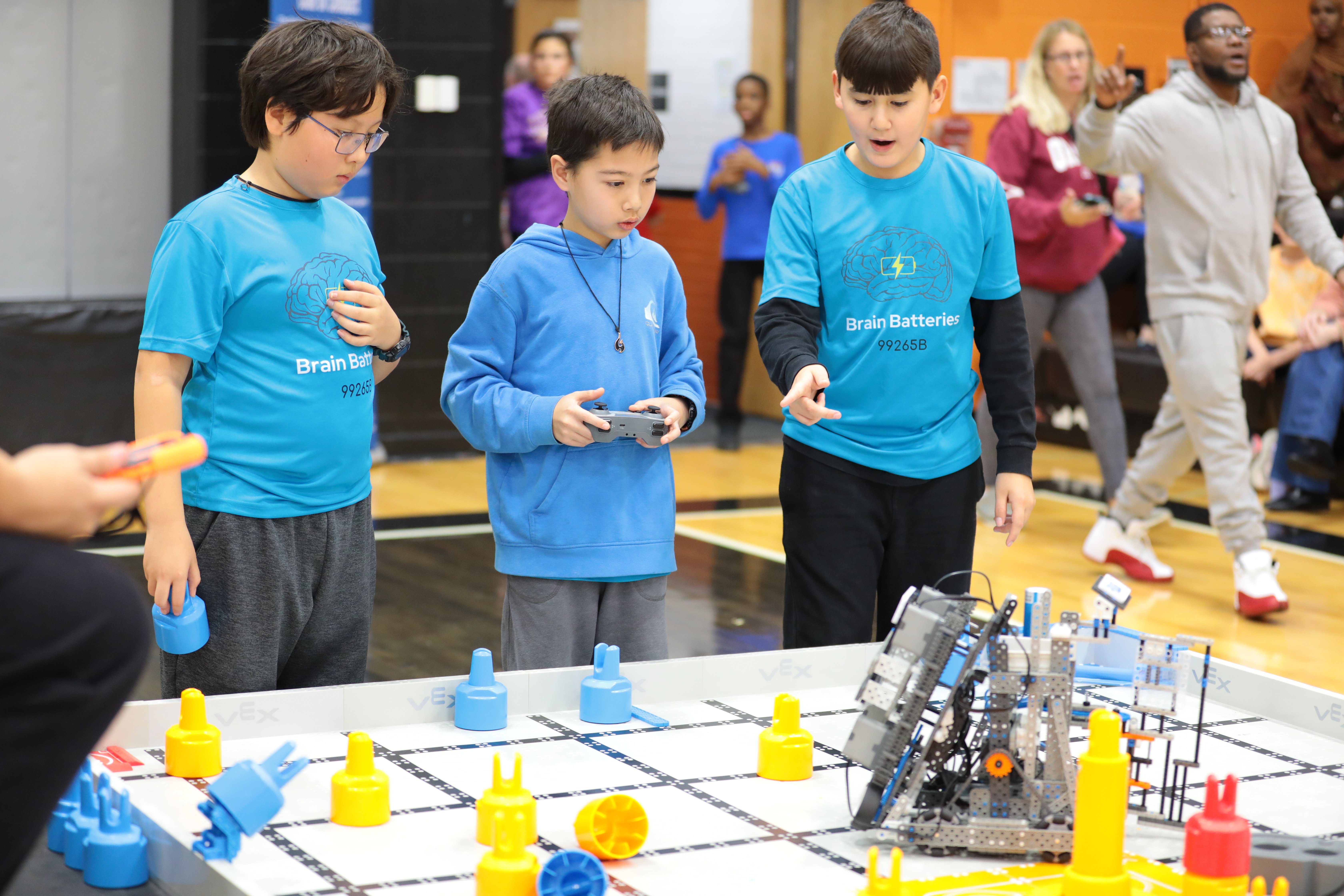 Three young students in matching teal team shirts operate a VEX IQ Robotics Competition robot on a competition field scattered with yellow, blue, and red game elements. One student holds a game controller while a teammate points toward the robot on the field.
