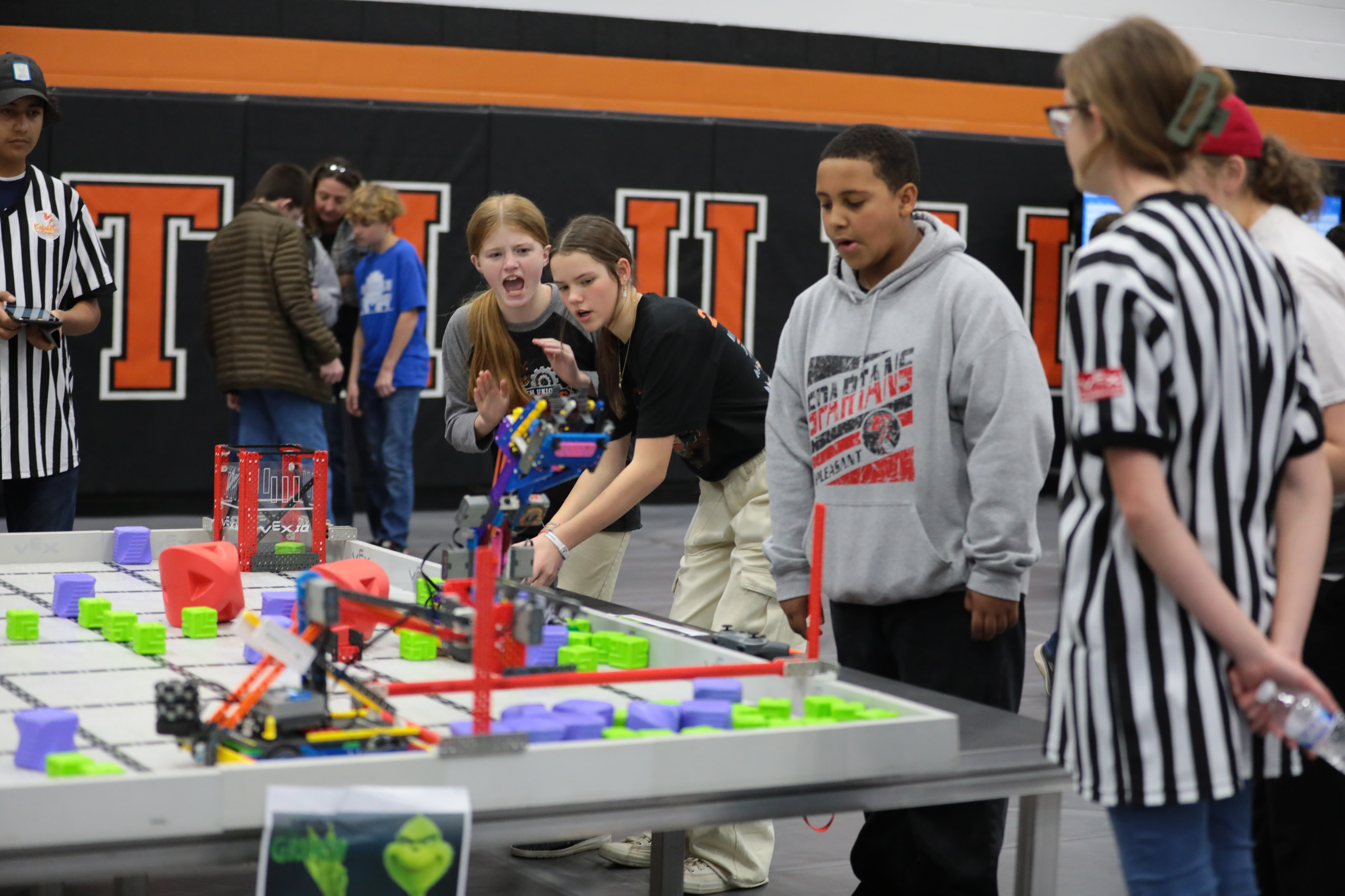 Two girls stand at a VEX IQ Field during a competition, yelling and focused on their robot's performance. Members of another team and a judge look on.
