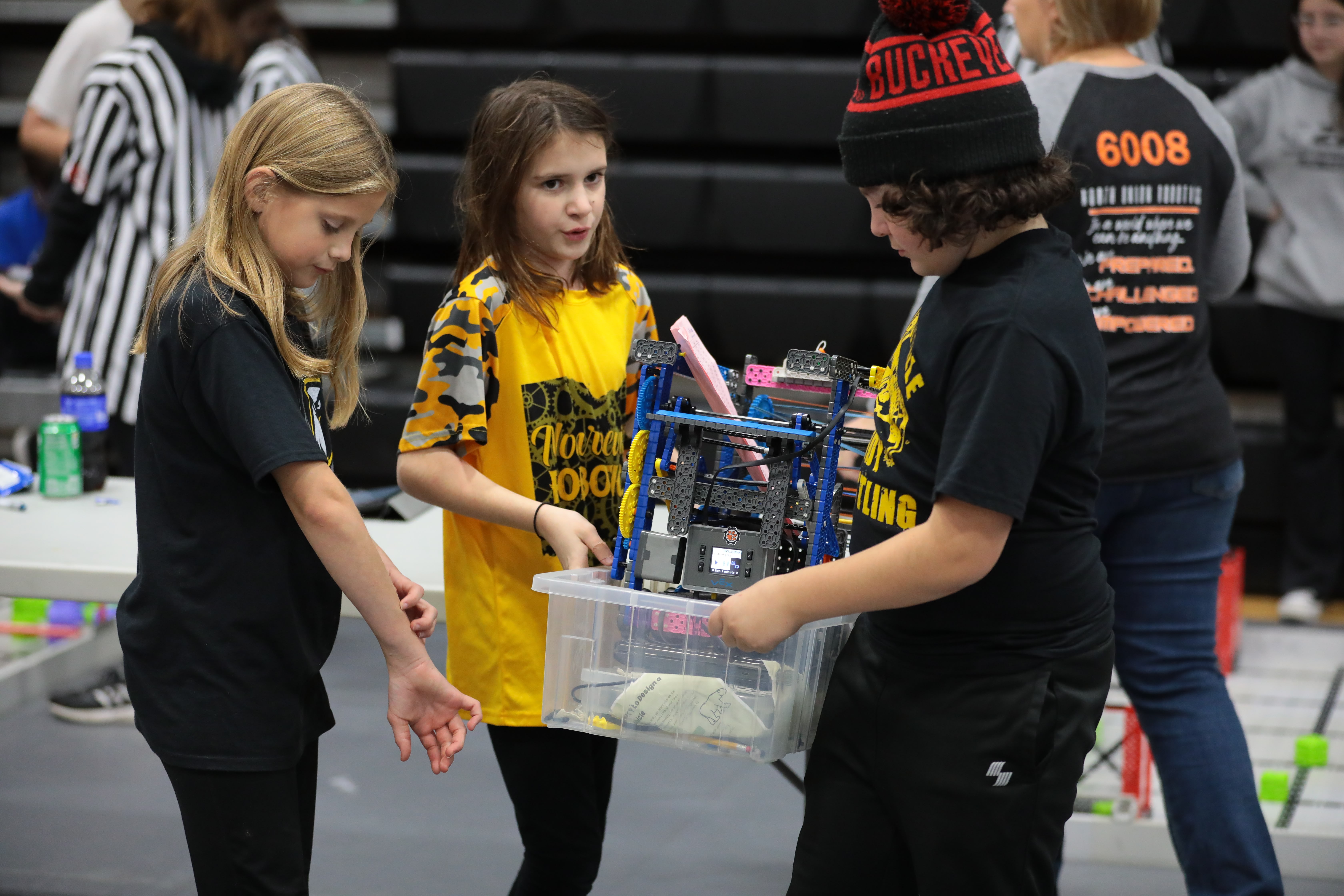 Three students carry their IQ Robot in a plastic container. Behind them is a set of bleachers and a person in a black and white striped referee jersey.