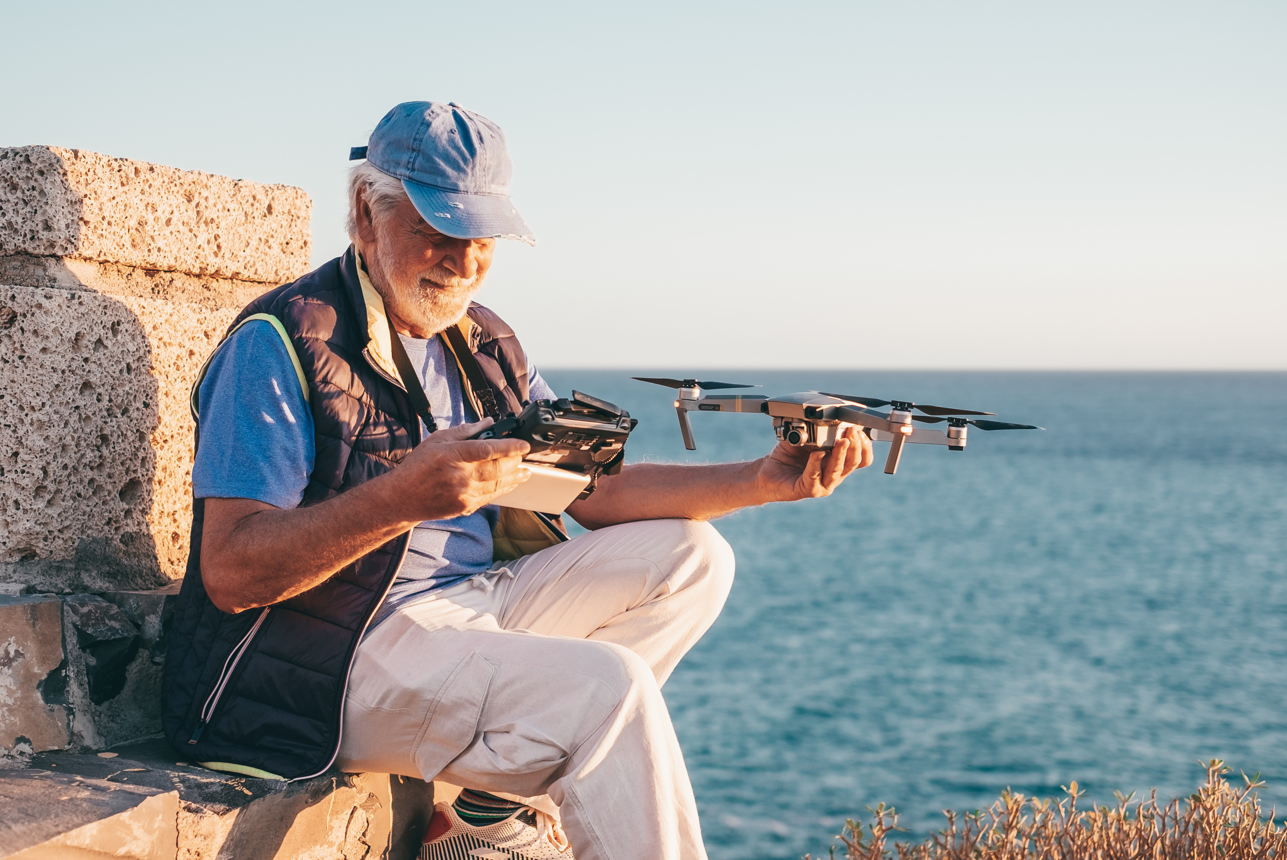 An older gentleman sits against a rock outcropping overlooking the ocean, holding a drone in one hand, and viewing video footage on a controller in the other hand.