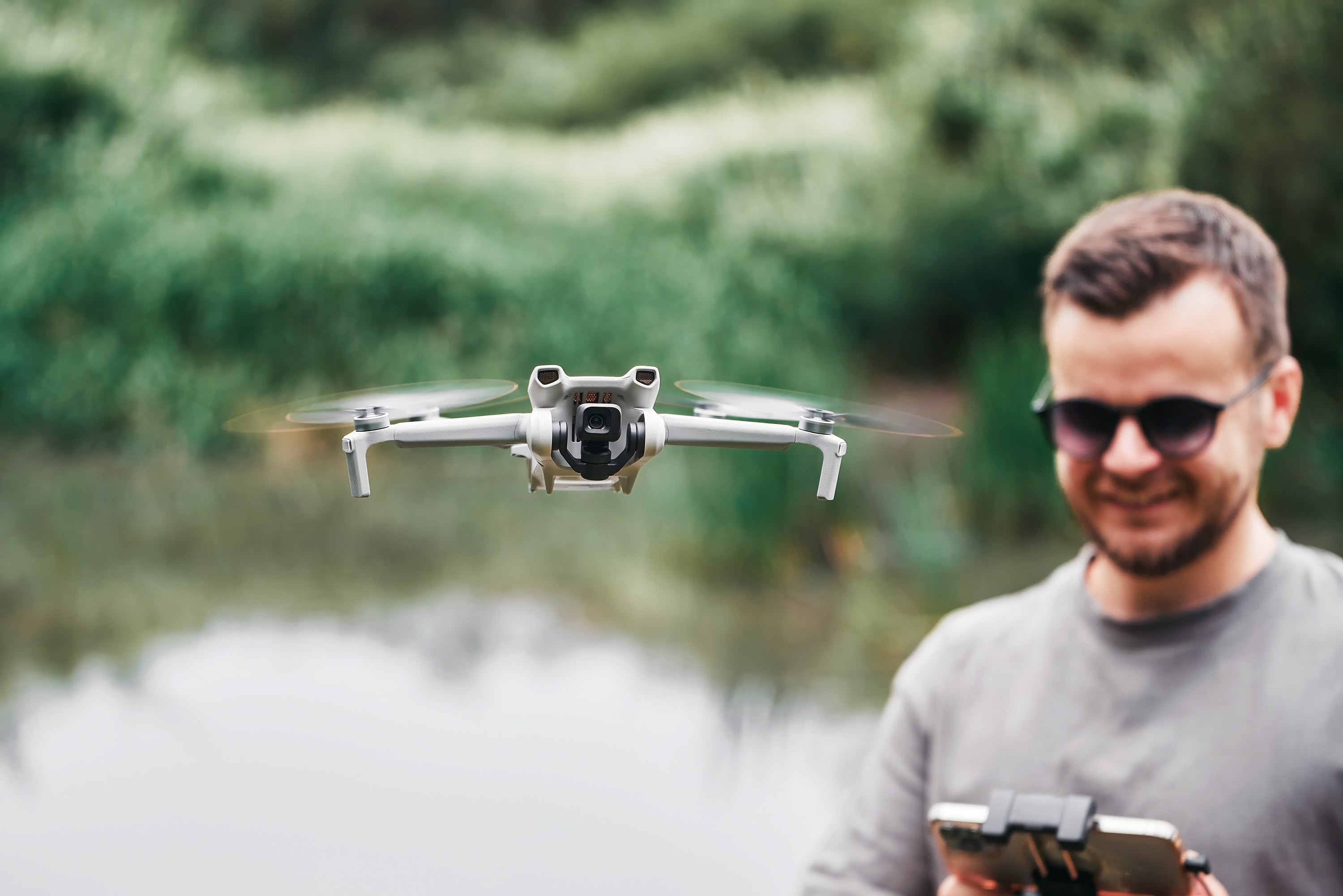 A man in sunglasses smiles while looking down at a drone controller in the background, while in the foreground, the drone flies above a wetland environment.
