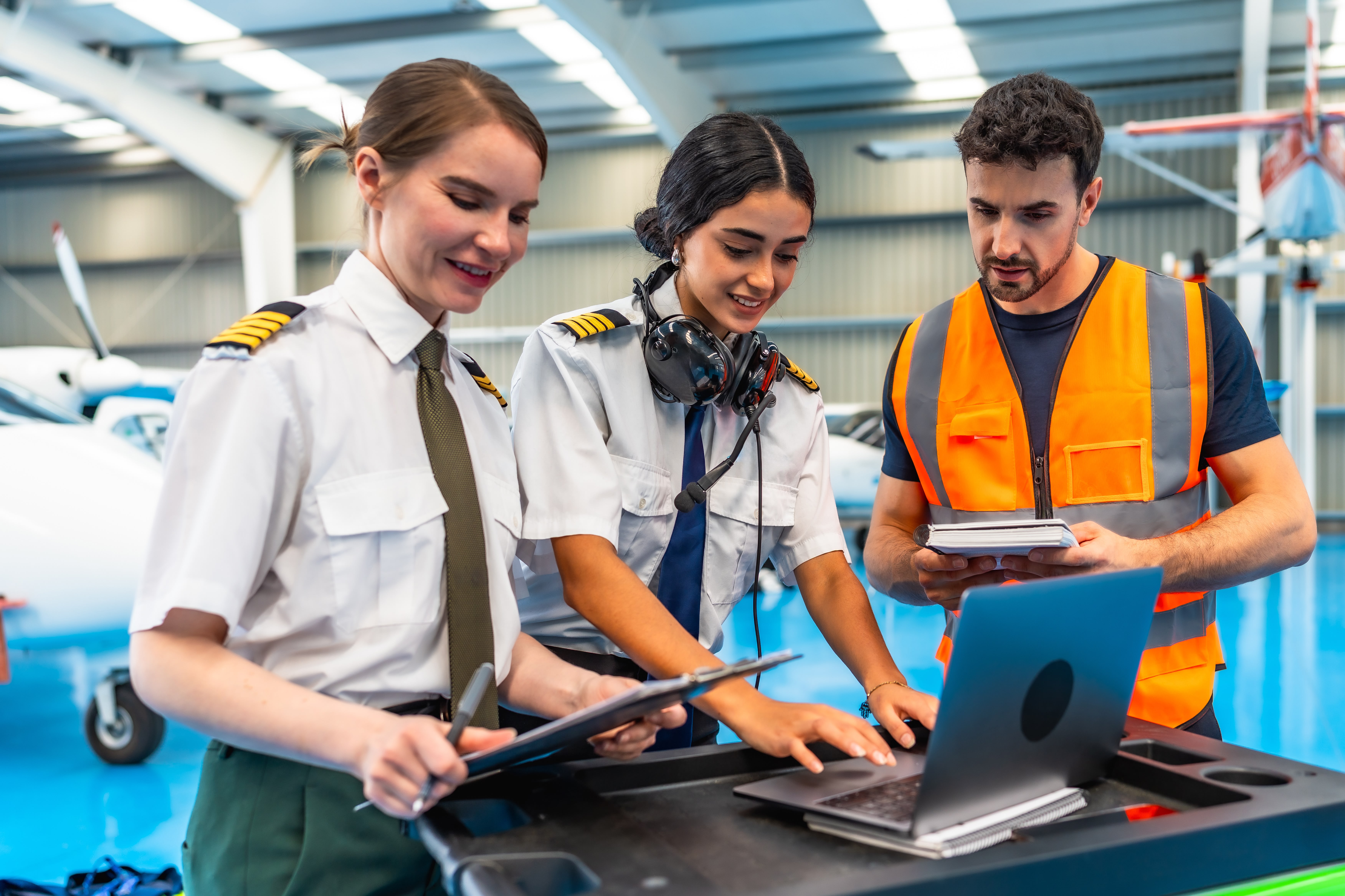 Two female pilots and a maintenance worker complete pre-flight checklist together with clipboards, laptops, and notebooks.