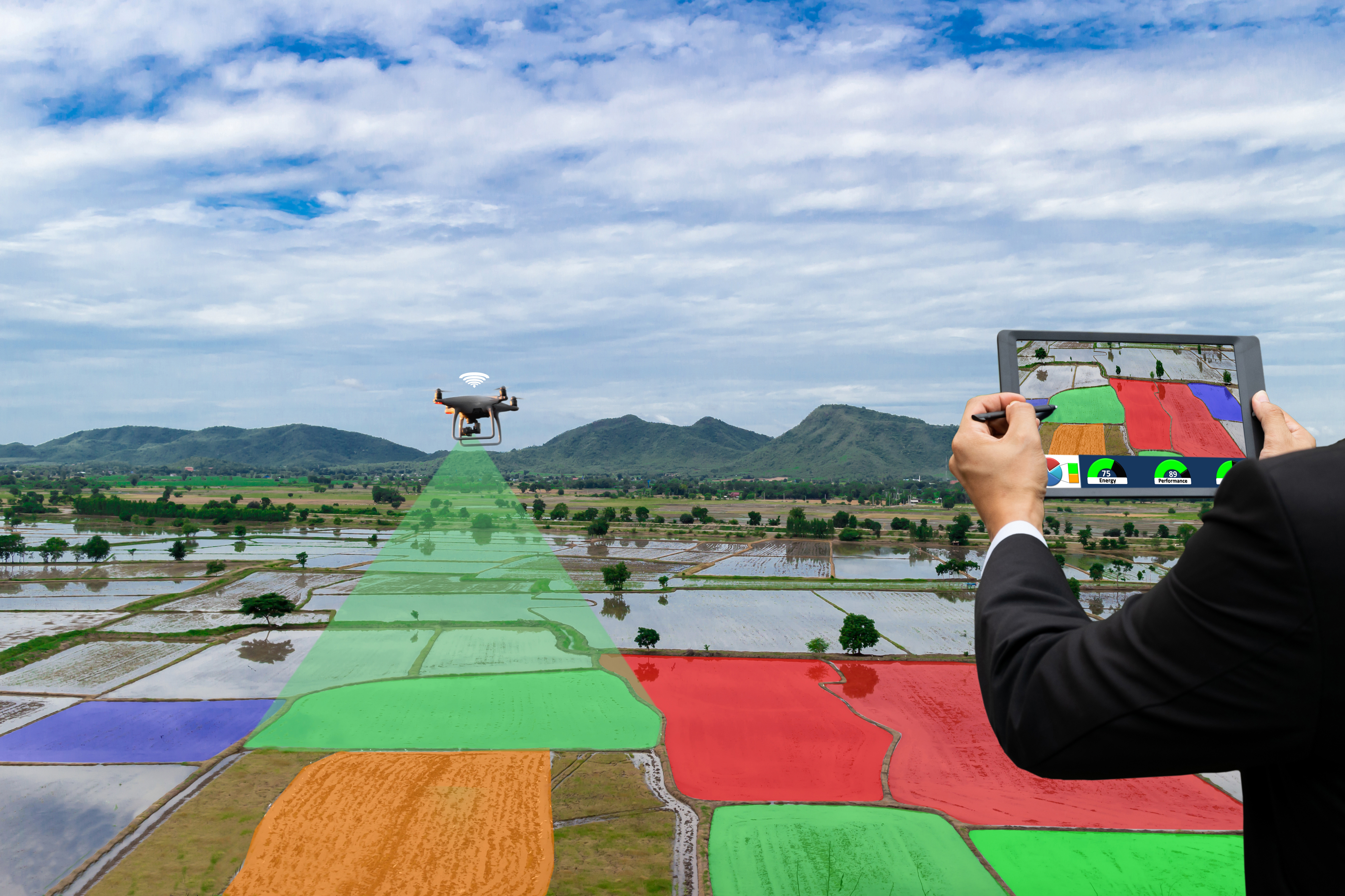 A drone flies above a field to collect data and visuals about crops being grown. A green cone indicates where the drone is getting data from, while in the foreground a person holds up a tablet showing the field and points to that area with a stylus.