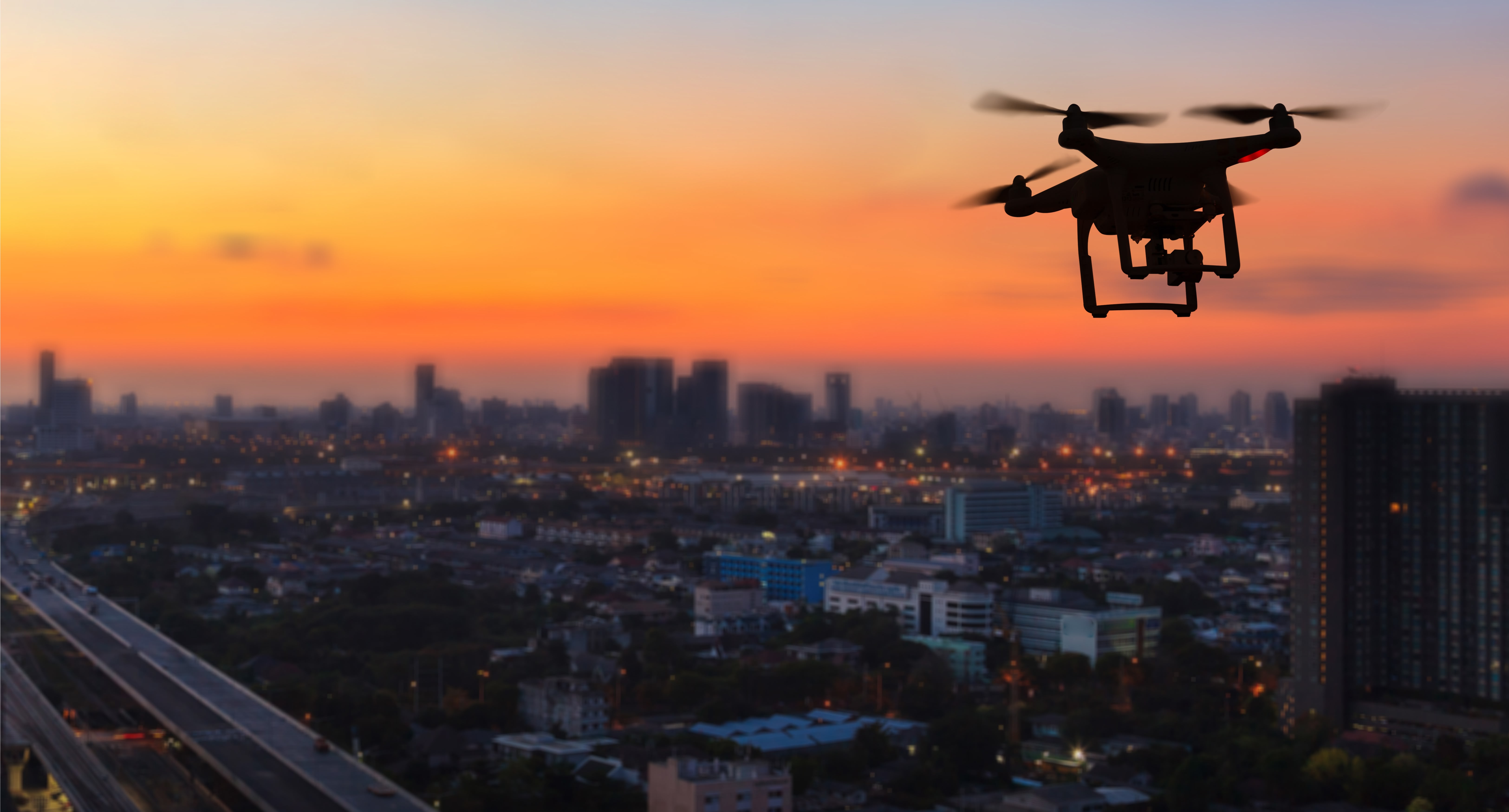 A drone flies into the sunset with a cityscape in the background.