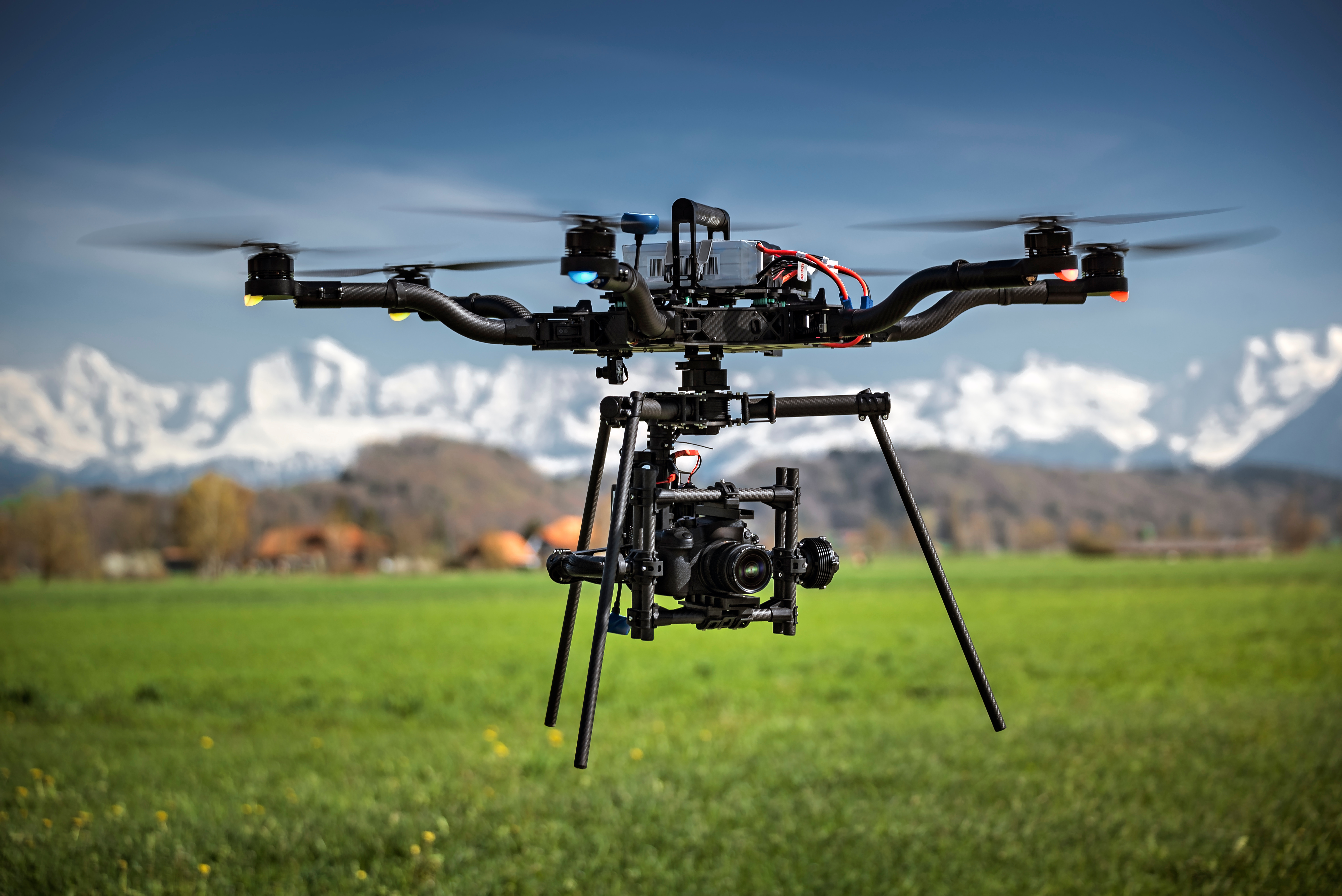 A drone flies over a field with a mountainous landscape in the background, with a hight tech camera attached to the base.