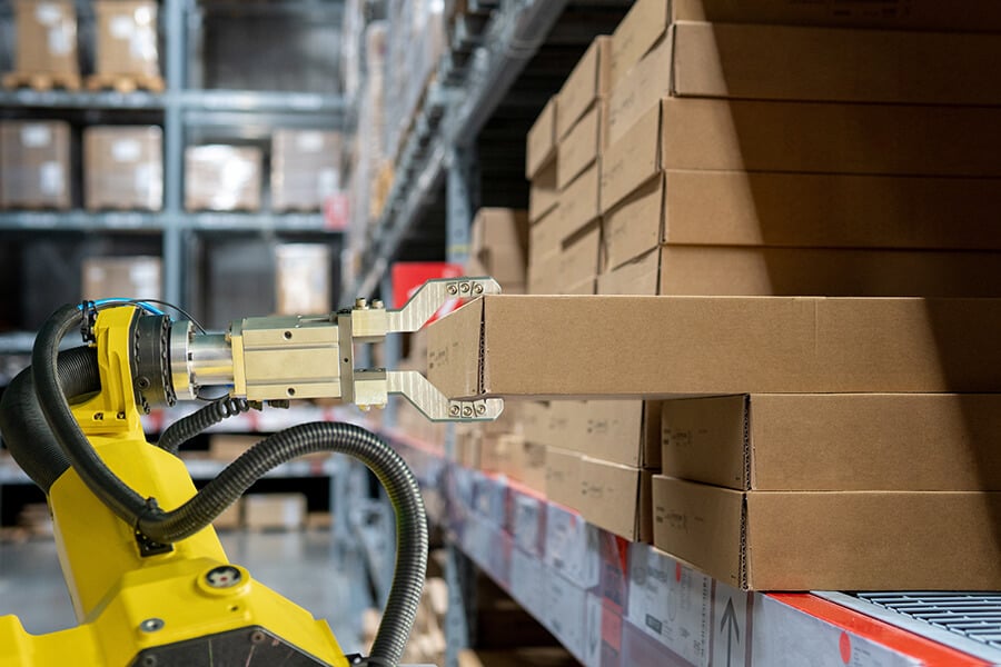 A robotic arm with a gripper attachment carefully lifting a cardboard box from a shelf in a warehouse. The shelf is stacked with uniform boxes, and the robotic arm’s precision demonstrates automated handling within the organized storage area.
