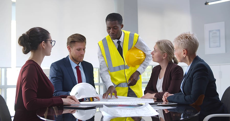 Five people are gathered around a table looking at blueprints. One man, wearing a yellow vest and holding a hard hat, is standing up, as if he is leading the discussion.