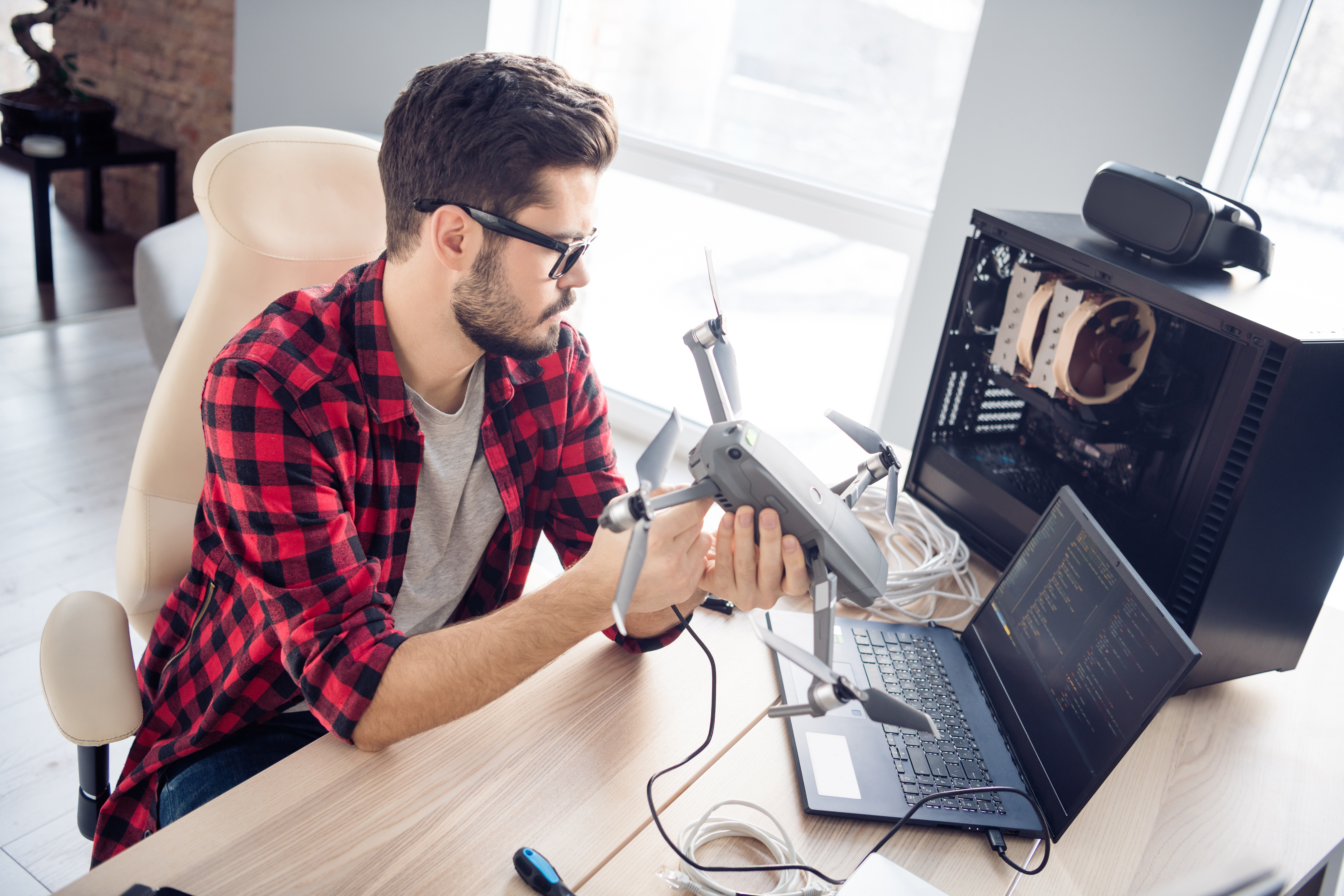 A man in a flannel shirt holds a drone in one hand as he connects a wire from a computer into the drone, while looking at drone data on the screen.