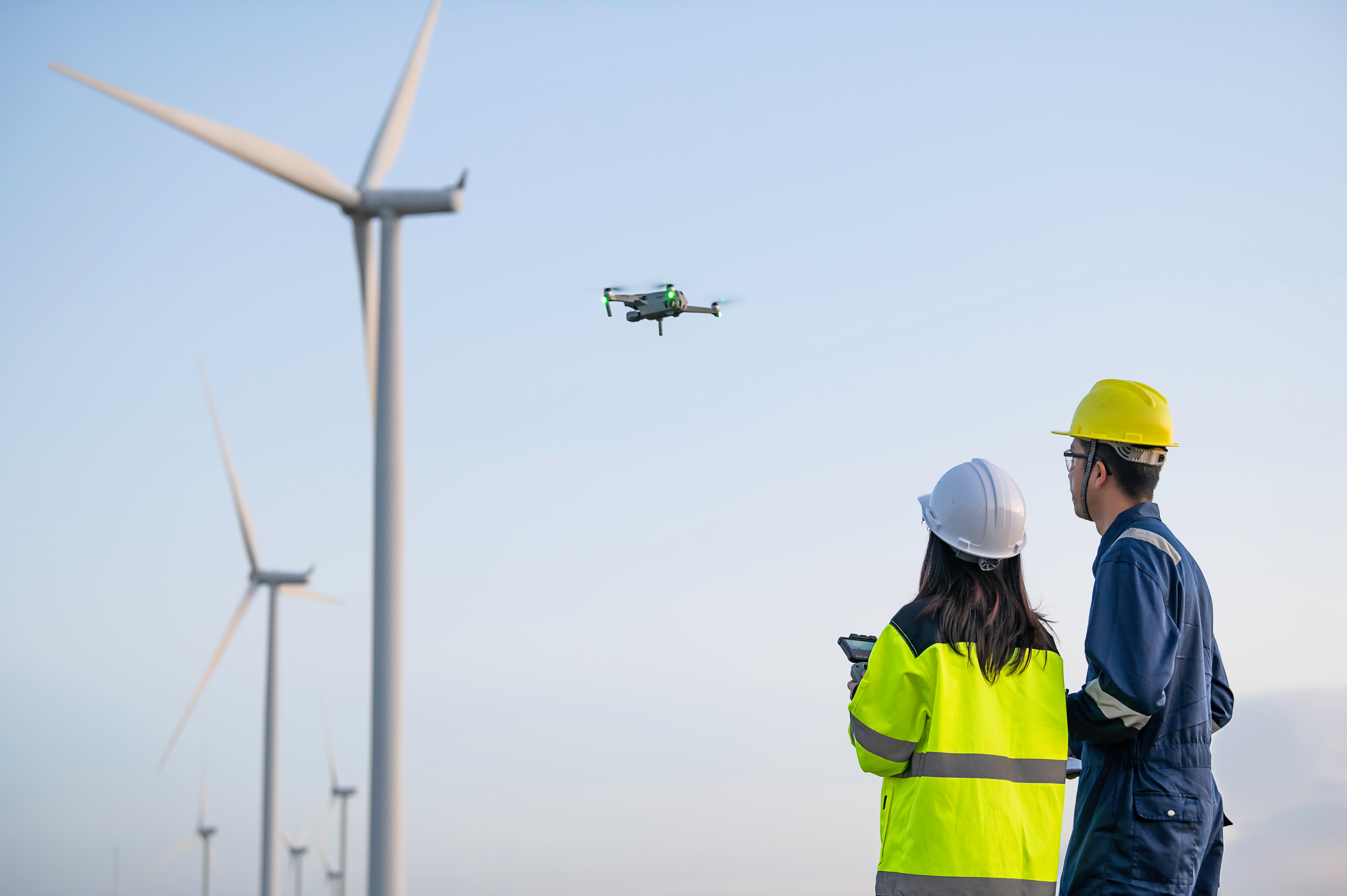 Two drone operators, both wearing hard hats and one holding a controller, watch a drone as it flies towards a line of wind turbines.