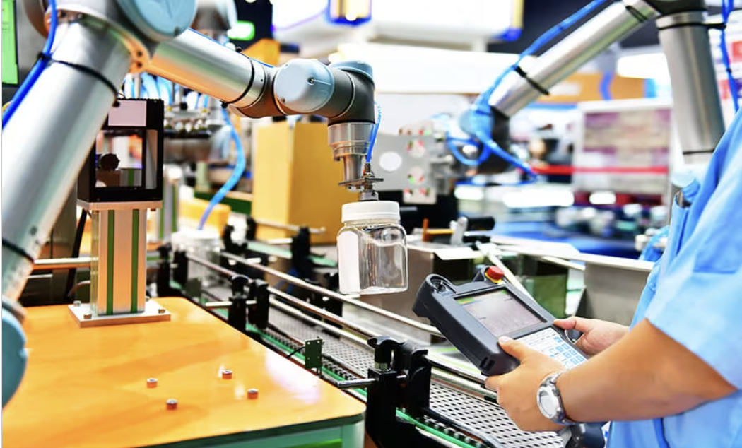 Un pendentif d'enseignement dans une usine tenu par un ouvrier. 2 bras robotisés sont en arrière-plan, manipulant des produits.