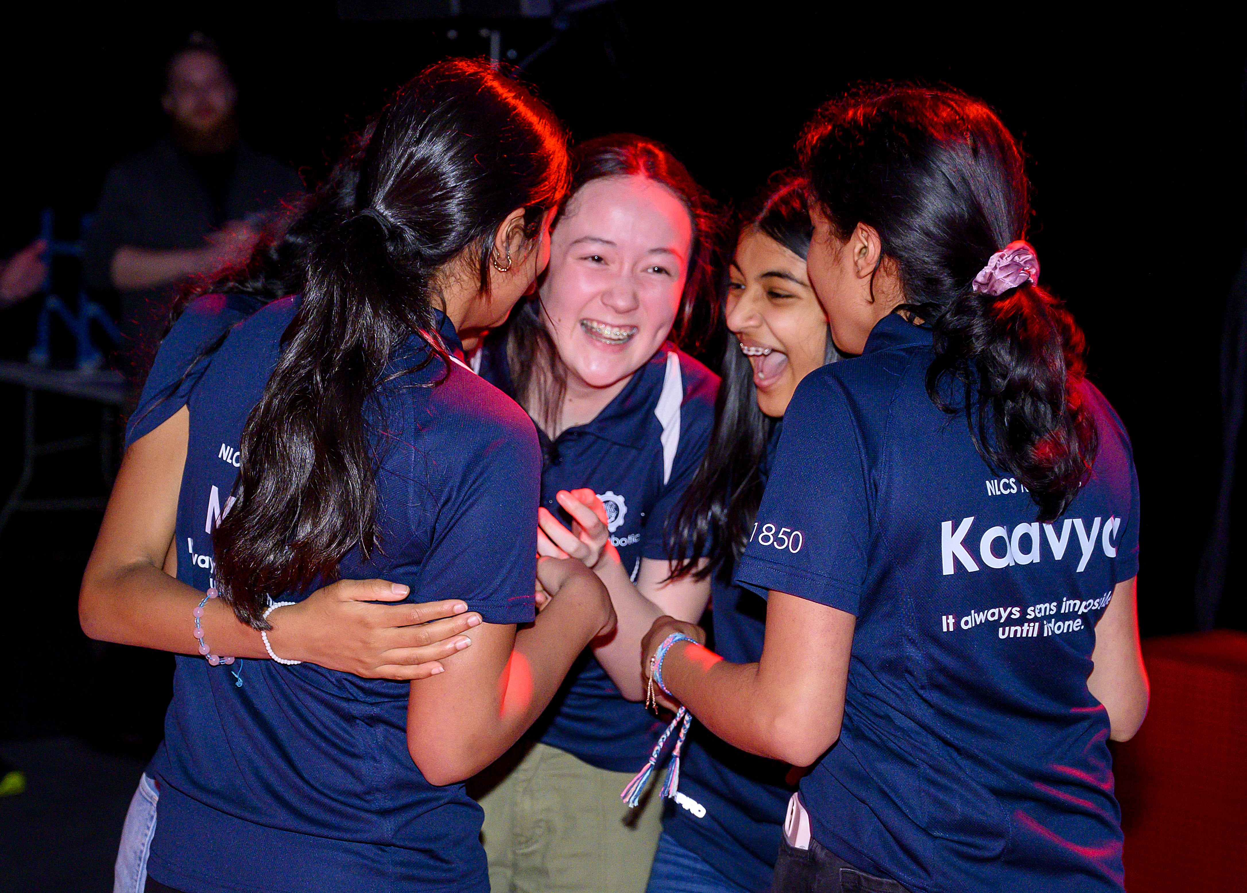 Four middle school aged girls in robotics team shirts hug each other in celebration.