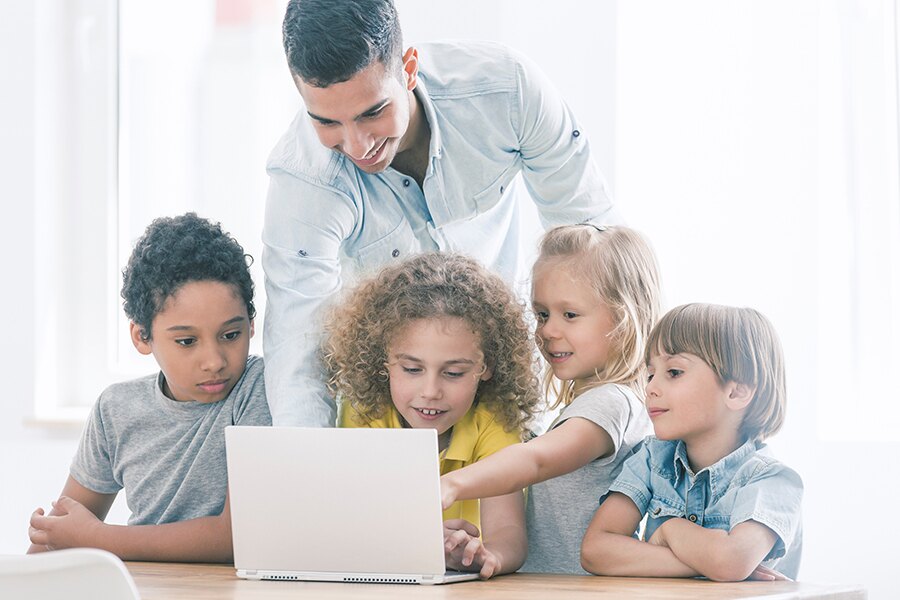 A group of students are gathered around a computer with their teacher behind them, working together to complete a coding project.