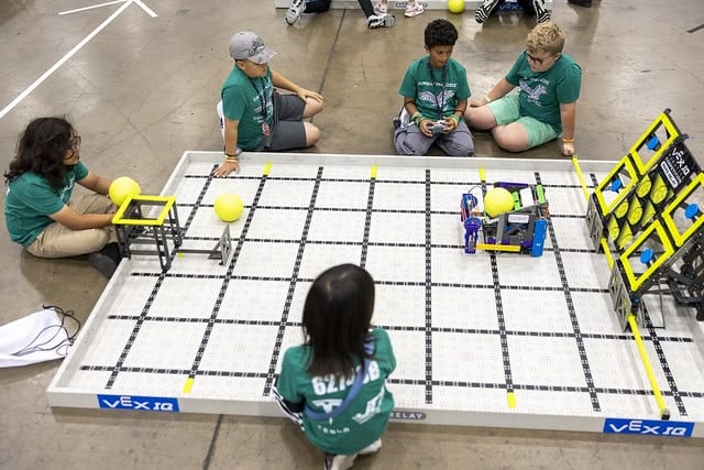 A team of 5 students sit around a practice field on the floor, while one student drives the robot. The other four look on to evaluate their driving strategy.