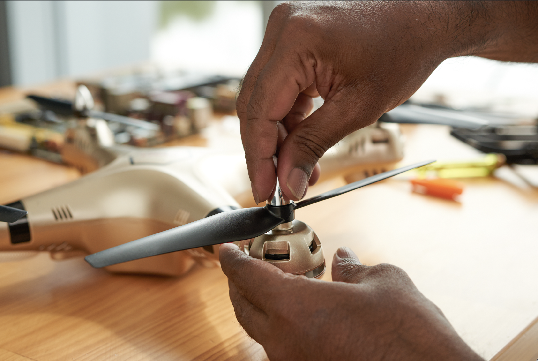Two hands hold a propeller mechanism that is detached from a drone on a table, to tighten the propeller blades.